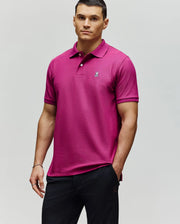 A man wears a Psycho Bunny MENS CLASSIC PIQUE POLO - B6K001F200 in bright magenta, featuring an embroidered Bunny logo on the chest, paired with black pants, against a plain light background.