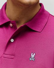 A close-up of a person wearing the Psycho Bunny MENS CLASSIC PIQUE POLO (B6K001F200) in magenta, made from soft Pima cotton, with white buttons and the embroidered Bunny logo with crossbones on the chest.