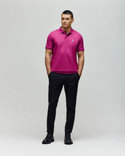 A man stands against a plain background wearing Psycho Bunny’s MENS CLASSIC PIQUE POLO - B6K001F200 in bright pink, featuring the embroidered Bunny logo, with black pants and shoes, hands in pockets and looking to the side.