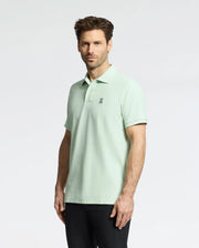 A man wears a Psycho Bunny men's classic piqué polo (B6K001F200) in light green, featuring a small embroidered Bunny logo on the left chest. His short brown hair pairs well with black pants against a plain light gray background.