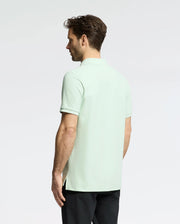 A man with short brown hair models the Psycho Bunny Men's Classic Piqué Polo (B6K001F200) in light green. Made from soft Pima cotton, it features a subtle bunny logo. He pairs it with black pants against a plain light gray background, facing away from the camera.