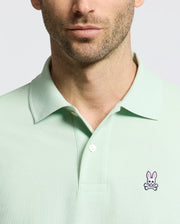 A man wears a Psycho Bunny MENS CLASSIC PIQUE POLO (B6K001F200) in light green, highlighted by its pink bunny and crossbones logo. Made from Pima cotton, it features a comfortable collar with two buttons undone against a plain white backdrop.
