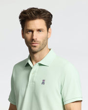 A man in a Psycho Bunny men's Classic piqué polo (B6K001F200), light mint green and made from soft Pima cotton, with a small embroidered Bunny logo on the chest, stands against a plain gray background with short brown hair and facial hair, looking directly at the camera.