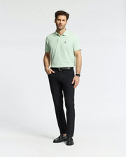 A person stands confidently against a plain background wearing a Psycho Bunny MENS CLASSIC PIQUE POLO (B6K001F200) in light green with the iconic Bunny logo, paired with black pants and shoes, one hand in their pocket.