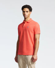 A person stands wearing the Psycho Bunny MENS CLASSIC PIQUE POLO - B6K001F200 in bright orange with an embroidered bunny logo on the chest, paired with light beige pants against a plain, light-colored background.