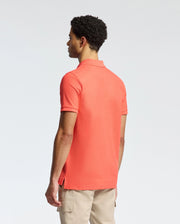 A man with short curly hair wears a Psycho Bunny coral MENS CLASSIC PIQUE POLO - B6K001F200, featuring a subtle bunny logo. He pairs it with beige pants and gazes left against a plain white background.