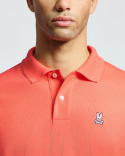 A man models the Psycho Bunny MENS CLASSIC PIQUE POLO - B6K001F200 in coral, made from soft Pima cotton. The upper torso shot showcases the small embroidered bunny with crossbones on the left chest.