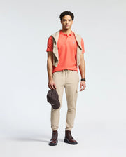 A man in a Psycho Bunny MENS CLASSIC PIQUE POLO - B6K001F200 with an embroidered Bunny logo, beige cargo joggers, and brown boots stands against a plain background. A beige Pima cotton sweater is draped over his shoulders as he holds a dark cap in his right hand.