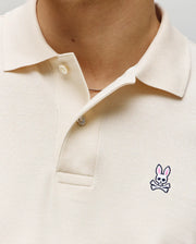 Close-up of a person wearing the Psycho Bunny MENS CLASSIC PIQUE POLO SHIRT - B6K001F200 in cream, featuring two buttons undone and an embroidered Bunny logo with pink ears and crossbones on the left chest.