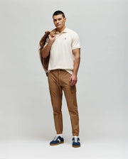 A man stands against a plain background wearing a cream Psycho Bunny MENS CLASSIC PIQUE POLO SHIRT (B6K001F200), tan cargo pants, blue-and-white sneakers, and holds a tan jacket over his left shoulder while looking forward neutrally.