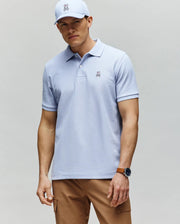 A man stands with his hands by his sides, wearing a Psycho Bunny MENS CLASSIC PIQUE POLO - B6K001F200 in light blue, paired with a matching bunny logo cap, tan pants, and a watch while gazing slightly toward the camera.