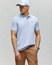 A man in a Psycho Bunny MENS CLASSIC PIQUE POLO - B6K001F200 in light blue with a small bunny logo, paired with a matching cap and tan cargo pants, stands against a gray background looking to the side.
