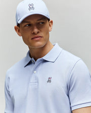 A man in a light blue Psycho Bunny MENS CLASSIC PIQUE POLO - B6K001F200 and matching cap, both featuring the signature bunny with crossed bones, looks off to the side against a plain background.