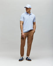 A man stands against a light gray background wearing a light blue Psycho Bunny MENS CLASSIC PIQUE POLO - B6K001F200, tan cargo pants, navy and white sneakers, and a matching light blue baseball cap.