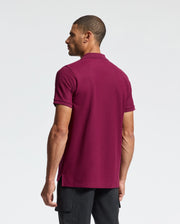 A man with short dark hair stands facing away. He's wearing a Psycho Bunny maroon MENS CLASSIC PIQUE POLO SHIRT, B6K001Z1PC, made of soft Pima cotton with an embroidered Bunny logo, paired with black pants against a plain white background.
