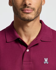 A person wears a Psycho Bunny MENS CLASSIC PIQUE POLO SHIRT (B6K001Z1PC) in maroon, featuring an embroidered Bunny logo with a cartoon skull and bunny ears on the chest. Crafted from luxurious Pima cotton, the focus is on the upper body and shirt against a plain light gray background.