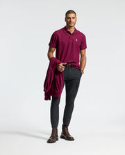 A man stands confidently against a plain background wearing Psycho Bunny's Men's Classic Piqué Polo Shirt - B6K001Z1PC with an embroidered Bunny logo. He pairs it with dark pants, brown boots, and has a maroon jacket draped over his left arm, showcasing the refined touch of Pima cotton in his attire.