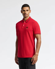 A person is seen wearing the Psycho Bunny MENS PIERCE PIQUE POLO - B6K201E200, a red polo shirt crafted from soft Pima cotton with black stripes detailing the collar and sleeves, standing against a plain background. This stylish polo, complete with an embroidered bunny logo on the chest, is complemented by sleek black pants.