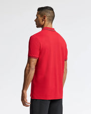 A man with short black hair stands facing away, wearing the Psycho Bunny Men's Pierce Pique Polo - B6K201E200, featuring a red Pima cotton design with black trim on the collar and sleeves. The embroidered Bunny adds a touch of charm to his look, which he effortlessly pairs with sleek black pants against a plain white backdrop.