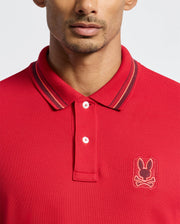 A person is dressed in the Psycho Bunny Men's Pierce Pique Polo (B6K201E200), which features a vibrant red color made from premium Pima cotton. The shirt is highlighted with dark striped detailing on the collar and adorned with an embroidered bunny skull logo on the chest. The polo also includes two fastened buttons, set against a simple, light-colored background.