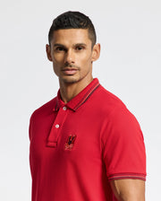 A man wearing the Psycho Bunny MENS PIERCE PIQUE POLO - B6K201E200, featuring a red Pima cotton fabric with an embroidered bunny emblem on the chest, stands against a plain background. His hair is short and styled, and he gazes directly at the camera.