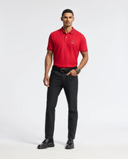 A man stands confidently in a red MENS PIERCE PIQUE POLO - B6K201E200 by Psycho Bunny, featuring an embroidered Bunny logo. He pairs it with black pants and shoes, complemented by a dark belt and watch for a sleek touch. His casual pose includes one hand in his pocket against a plain white background.