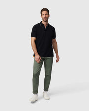 A man with short hair and a beard stands against a plain background. He is wearing an olive green Psycho Bunny MENS EAST HILLS JOHNNY COLLAR POLO SHIRT - B6K331B200 made of Pima cotton, paired with pants and white sneakers. He has a relaxed expression and one hand slightly extended, with the other by his side.