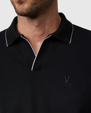 A close-up view of a person wearing a Psycho Bunny MENS EAST HILLS JOHNNY COLLAR POLO SHIRT - B6K331B200 with white trim on the Johnny collar and a small embroidered logo of a rabbit with crossbones on the left chest. The background is plain and light-colored.