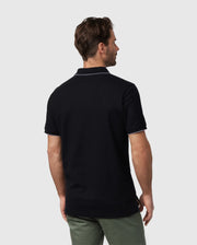 A man with dark hair stands with his back to the camera, wearing a black Psycho Bunny MENS EAST HILLS JOHNNY COLLAR POLO SHIRT - B6K331B200 with white trim on the Johnny collar and sleeves. He also wears green pants. The background is plain and light-colored.