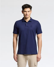 A man stands against a plain background wearing the Psycho Bunny MENS NICHOLSON RIB COLLAR PIQUE POLO in navy, crafted from luxurious Pima cotton, and beige pants. The shirt features an embroidered logo on the chest. He has short dark hair and a neutral expression.