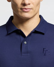 A person wearing Psycho Bunny's dark blue polo, the Nicholson Rib Collar Pique Polo - B6K401F200, made from premium Pima cotton with a small embroidered logo on the chest. The focus is on the upper torso, emphasizing the collar and buttons against a light background.