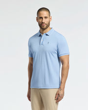 A bearded man wears the Psycho Bunny Men's Bryce Performance Pique Polo in light blue, featuring a small left-side logo, and beige pants as he stands against a white background, highlighting the breathable fabric.