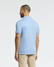 A man faces away from the camera in a light blue Psycho Bunny Bryce Performance Pique Polo (B6K486F200) and beige pants. This quick-dry shirt, made from breathable performance pique fabric, is set against a simple light grey background.