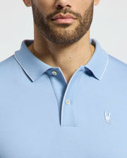 A person wearing the Psycho Bunny Men's Bryce Performance Pique Polo, B6K486F200, in light blue with a small embroidered logo on the chest. The shirt has a classic collar, button placket, and is made of breathable performance pique against a plain white background.