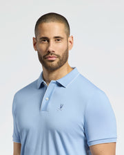 A man with short hair and a beard wears a Psycho Bunny MENS BRYCE PERFORMANCE PIQUE POLO - B6K486F200 in light blue, featuring breathable fabric and a small embroidered logo on the chest. He looks directly at the camera against a plain, light background.