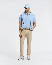 A man wearing a light blue Psycho Bunny Bryce Performance Pique Polo, matching baseball cap, and beige pants stands against a simple background. He has a light blue sweater draped over his shoulders and is wearing quick-dry white sneakers.