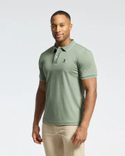 A man stands against a plain background wearing a light green Psycho Bunny Men's Bryce Performance Pique Polo, B6K486F200, with breathable moisture-wicking fabric and a small chest logo. He's also in beige pants, with a neutral expression and no other objects in view.