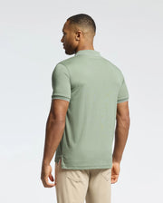 A man with short hair stands facing away, wearing the Psycho Bunny MENS BRYCE PERFORMANCE PIQUE POLO in light green, crafted from breathable, moisture-wicking fabric, paired with beige pants. The white-trimmed shirt aligns well with a plain white background.
