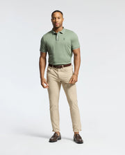 A confident man wears a Psycho Bunny MENS BRYCE PERFORMANCE PIQUE POLO in green, crafted from breathable, moisture-wicking fabric. He pairs it with beige pants, a brown belt, and shoes, exuding relaxed elegance with his short haircut against a plain white backdrop.