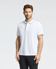A man with short brown hair and a beard sports a Psycho Bunny MENS BRYCE PERFORMANCE PIQUE POLO (B6K486F200) in white with black accents, paired with black pants. He stands against a plain light gray background, embodying comfort and style.