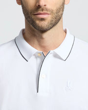 A man wears the MENS BRYCE PERFORMANCE PIQUE POLO by Psycho Bunny, featuring a white design with black trim on the collar and a rabbit logo. Made from Performance Pique Fabric, this stylish polo offers comfort with its moisture-wicking qualities.