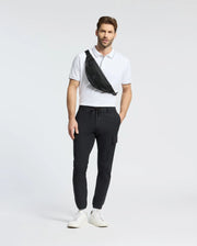 A man stands against a plain background wearing a Psycho Bunny MENS BRYCE PERFORMANCE PIQUE POLO in white, black jogger pants, and white sneakers. He carries a black crossbody bag slung over his shoulder.