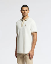 A man stands facing forward against a white background, wearing the Psycho Bunny MENS NEW CASTLE PIQUE POLO - B6K487F200 in slim fit, short-sleeved off-white style with beige collar, paired with beige shorts.