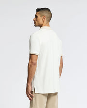 A man with short dark hair, standing and facing away from the camera against a plain light background, is wearing a white Psycho Bunny MENS NEW CASTLE PIQUE POLO - B6K487F200 and beige shorts.