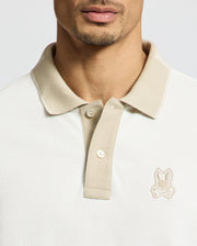 Close-up of a man wearing the Psycho Bunny MENS NEW CASTLE PIQUE POLO (B6K487F200), a slim fit light beige Pima Cotton polo with contrast collar detailing and a rabbit logo on the left chest, showing his neck, chin, and jawline.