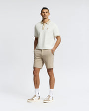 A man stands against a plain white background wearing the Psycho Bunny MENS NEW CASTLE PIQUE POLO - B6K487F200 in cream, paired with beige shorts, white socks, and beige sneakers. His hands are in his pockets as he looks at the camera.