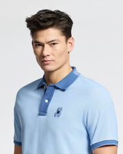 A person with short, dark hair wears a slim fit, light blue MENS NEW CASTLE PIQUE POLO - B6K487F200 from Psycho Bunny. Made from soft Pima cotton, it features a blue collar and an embroidered bunny patch on the chest. They stand against a plain white background, gazing into the camera.