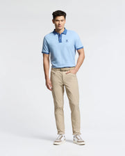 Wearing the MENS NEW CASTLE PIQUE POLO by Psycho Bunny, featuring a navy collar and embroidered chest Bunny patch, paired with khaki pants and white shoes with brown accents, a person stands against a plain background with one hand in their pocket.