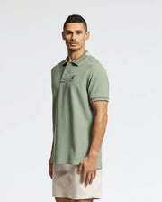 A man models a Psycho Bunny Men's New Castle Pique Polo (B6K487F200) in light green, crafted from soft Pima Cotton, with luxury finishes and an embroidered chest logo. He wears beige shorts and stands against a white background, facing the camera.