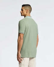 A man gazes sideways, wearing a Psycho Bunny MENS NEW CASTLE PIQUE POLO (B6K487F200) in rich Pima Cotton with beige pants, set against a plain white background that highlights the luxury details of his outfit.